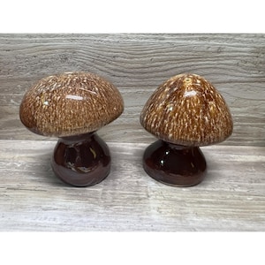 May include: Two ceramic mushroom-shaped decorative items. The mushroom caps are brown with white speckles, and the stems are a darker brown. The items are on a light wood-grain surface. The mushrooms are likely for home decor.
