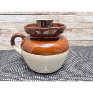 May include: A two-toned ceramic crock featuring a glossy dark brown upper section and a smooth cream-colored lower base. The vessel includes a matching dark brown lid with a central knob handle and a single curved side handle for easy gripping.
