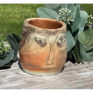 May include: A tan and terracotta colored ceramic planter featuring a hand-carved human face design. The vessel has a wide circular opening, a smooth interior, and textured details around the eyes, nose, and mouth etched into the clay surface.