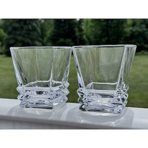 May include: Two clear glass tumblers featuring a square base that transitions into a wider, faceted rim. The heavy-bottomed drinking glasses have a geometric, stepped design at the base with multiple horizontal ridges and smooth, transparent walls.