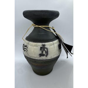 May include: A handcrafted ceramic vase with a black top and base, and a cream-colored band featuring embossed symbols. The vase is tied with a natural fiber string and has a tag attached. The vase is a decorative item.