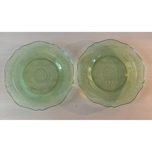 May include: Two light green glass plates with a scalloped edge and embossed floral design. The plates have a central medallion design and a decorative border. The plates are displayed on a white surface.