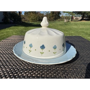 May include: A ceramic butter dish with a cream-colored, fluted dome lid featuring a hand-painted pattern of small blue flowers and green stems. The base is a light blue plate with a scalloped edge, providing a contrast to the speckled dome.