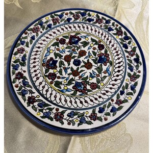 May include: A round, ceramic plate with a hand-painted floral design. The plate features a white background with a central pattern of blue, brown, and burgundy flowers and green leaves. A blue border surrounds the design, with a decorative white and blue outer edge.