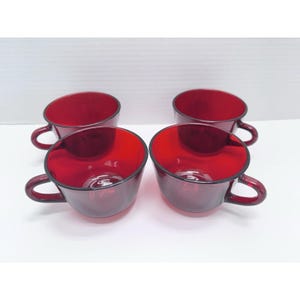 May include: A set of four matching ruby red glass cups, each featuring a smooth, rounded bowl shape and a curved handle on the side. The translucent red glass is uniform in color, with a polished finish and a simple, functional design.