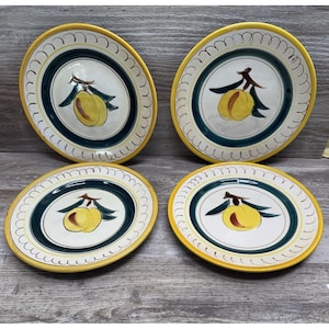May include: Four ceramic plates with a yellow rim and a central design featuring a yellow fruit with green leaves. The plates have a white background with a dark green and cream border.