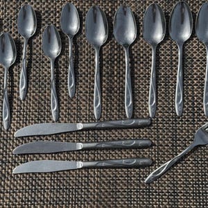 May include: A collection of stainless steel silverware, including spoons, knives, and a fork, arranged on a textured brown surface. The spoons have a teardrop shape and decorative handles. The knives and fork also have decorative handles.