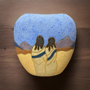 May include: A rounded, flat decorative stone featuring a painted landscape scene. Two figures with dark hair stand in the foreground, facing a desert horizon with yellow sand, brown hills, purple mountains, and a textured light blue sky above.