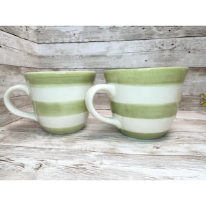 May include: Two ceramic mugs featuring horizontal stripes in light green and white. Each mug has a curved handle on the side. The mugs are positioned side by side, showcasing a consistent hand-painted appearance with a glossy finish on the exterior.