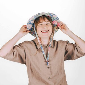 May include: A bucket hat featuring a blue background with a multi-coloured floral pattern of pink, yellow, and green flowers. The hat includes a matching fabric chin strap with a small brown wooden bead toggle for adjustment and a wide brim.