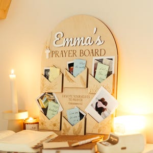 Puede incluir: Un tablero de oración de madera con el nombre "Emma's" y las palabras "Prayer Board" en escritura blanca. El tablero tiene varios pequeños bolsillos en forma de sobre que contienen notas y fotos. Una vela encendida y un libro abierto están sobre la mesa.