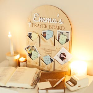 Op de afbeelding: Een boogvormig houten gebedsbord met de tekst Emma's PRAYER BOARD en DEVOTE YOURSELVES TO PRAYER Colossians 4:2. Het bord heeft zes houten envelopvormige vakjes met kleine kaartjes en foto's, versierd met gegraveerde bloemmotieven.