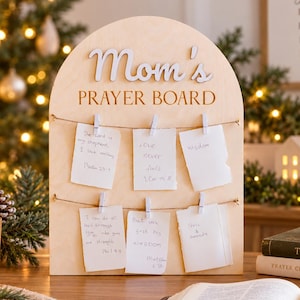 Puede incluir: Un tablero de oración de madera clara con forma arqueada y el texto Mom's PRAYER BOARD en letra cursiva y de molde. Dos cuerdas horizontales sostienen seis notas de papel blanco sujetas con pinzas de madera, con peticiones de oración.