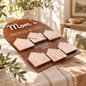 Puede incluir: Un tablero de oración de madera arqueado con acabado marrón oscuro que presenta el texto "Mom's PRAYER BOARD" y "DEVOTE YOURSELVES TO PRAYER Colossians 4:2". Seis bolsillos de madera clara en forma de sobre con diseños botánicos grabados.