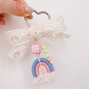 May include: A handmade keychain featuring a heart-shaped silver clasp, a cream-colored lace bow, and a rainbow macrame design. The keychain includes a pink gingham bead and colorful letter beads spelling "JEAN".