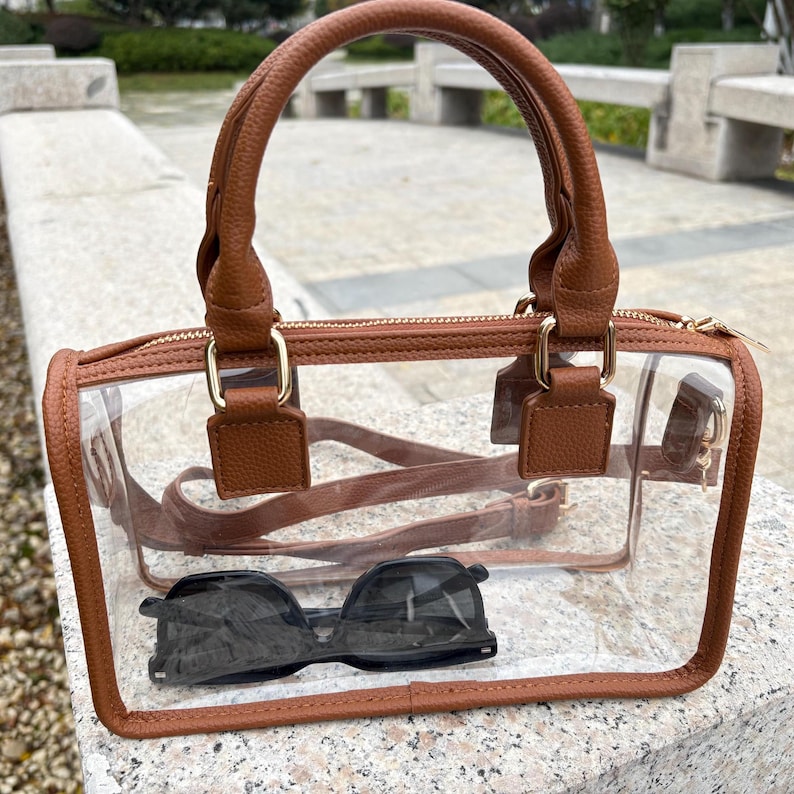 Transparent PVC stadium purse with brown trim displayed on a stone bench with sunglasses inside