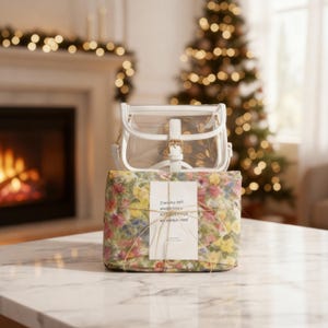 May include: A clear, white, and gold-toned handbag sits on a floral gift box tied with twine. The gift box has a tag that reads "Everyday isn't always happy, but happy things are always here!" A Christmas tree and fireplace are in the background.