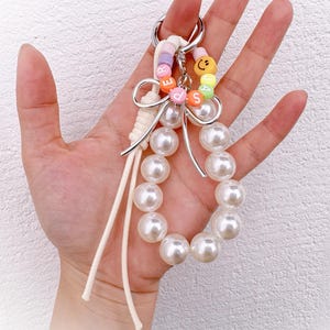 May include: A pearl beaded keychain with a silver-tone metal ring. The keychain features a loop of large, white faux pearls and a smaller loop with colorful letter beads spelling "BEADS" and a smiley face charm. White cord and a bow accent the design.