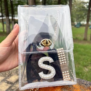 May include: A clear, rectangular crossbody bag with a gold chain strap and clasp. The bag contains a black item, a lipstick, and a black rectangle with a pearl-studded letter "S".