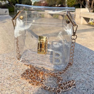 Peut inclure: Un sac à main transparent et clair avec une bandoulière et des accents dorés. Le sac est doté d'un fermoir doré et d'une lettre "B" décorative ornée de petites pierres transparentes. La chaîne est de couleur or chaud.