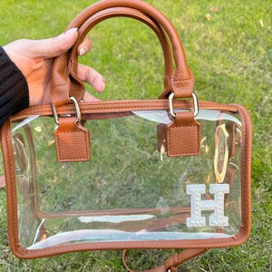 Brown clear stadium bag held outdoors with a sparkling chenille letter patch ‘H’ on the bottom corner.