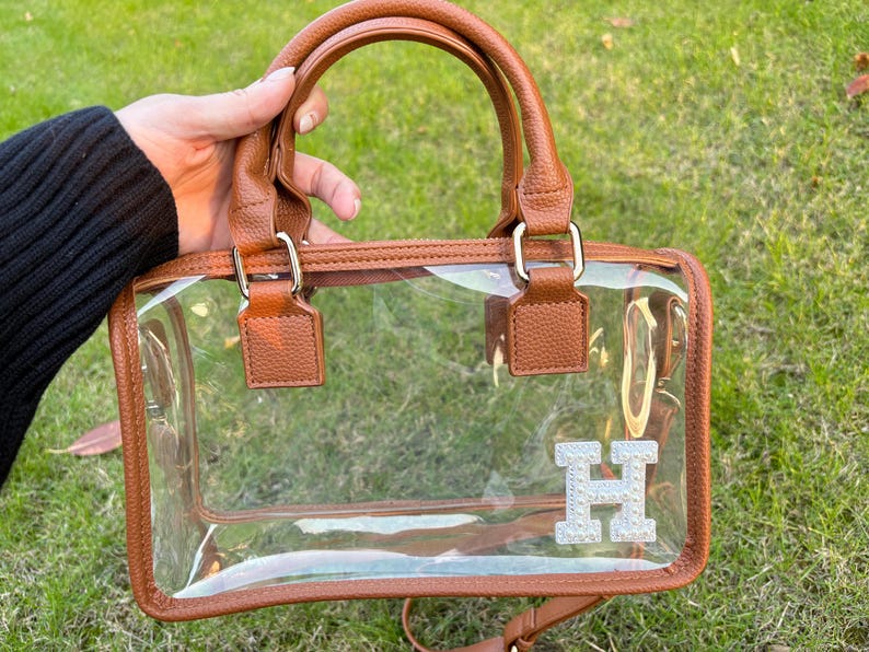 Brown clear stadium bag held outdoors with a sparkling chenille letter patch ‘H’ on the bottom corner.