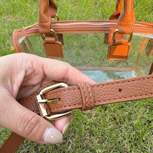 Close-up of adjustable brown strap and gold buckle of a clear stadium-approved handbag