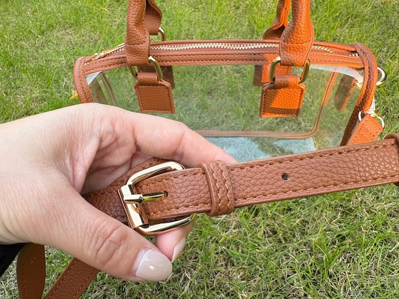 Close-up of adjustable brown strap and gold buckle of a clear stadium-approved handbag