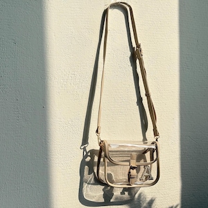 Può includere: Una borsa a tracolla trasparente con finiture e cinturino beige è appesa a una parete strutturata. La borsa presenta una chiusura con fibbia e una lettera decorativa "A" sul davanti. La borsa è sospesa da una sottile tracolla regolabile.
