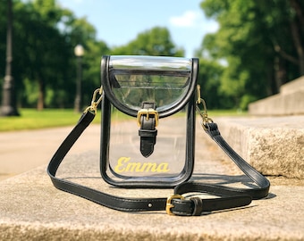 Può includere: Borsa a tracolla trasparente con finiture nere, una tracolla nera e accessori color oro. La borsa ha una chiusura con patta e una fibbia. Il nome "Emma" è stampato in giallo sul davanti. La borsa è su una superficie di pietra.