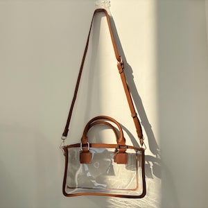 Clear crossbody stadium bag with long adjustable strap hanging on a wall under natural sunlight