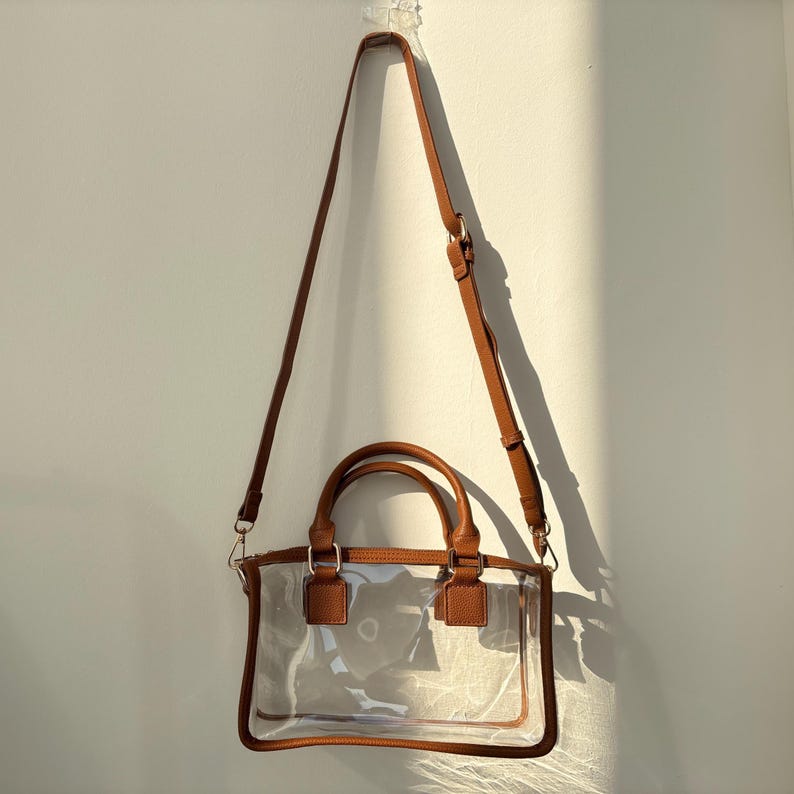 Clear crossbody stadium bag with long adjustable strap hanging on a wall under natural sunlight