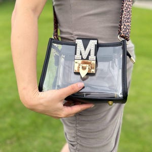 Puede incluir: Un bolso rectangular transparente con ribetes negros y un cierre dorado. El bolso presenta una letra "M" decorativa plateada y una correa con estampado de leopardo. El bolso se sostiene sobre un fondo verde.