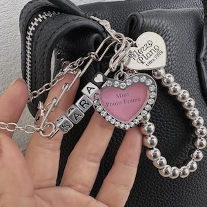 May include: A silver keychain with a heart-shaped mini photo frame, a heart-shaped charm with the text "Mezzo Piano Since 1988", and lettered beads spelling "SARA". The photo frame is surrounded by rhinestones.
