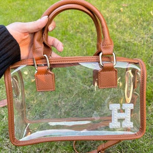 Brown clear stadium bag held outdoors with a sparkling chenille letter patch ‘H’ on the bottom corner.