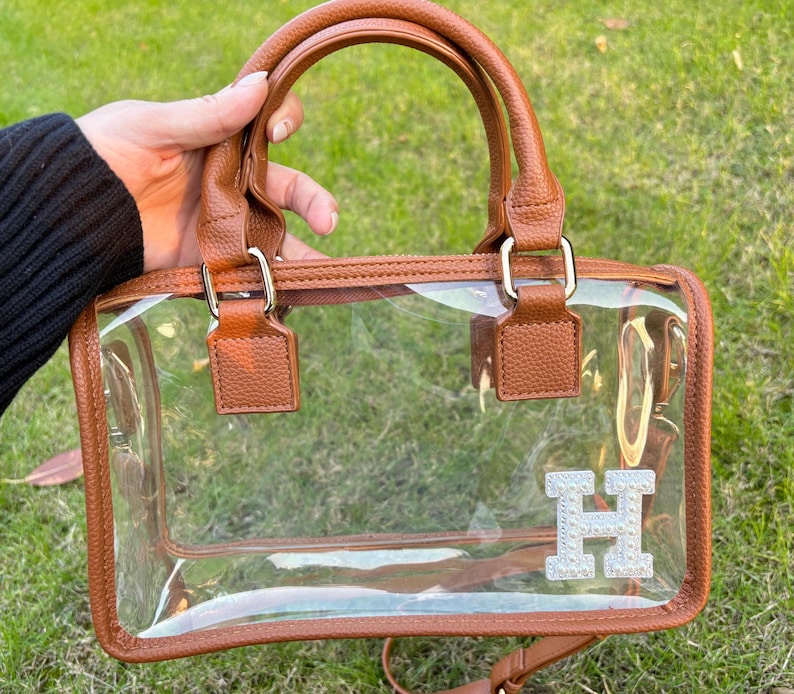 Brown clear stadium bag held outdoors with a sparkling chenille letter patch ‘H’ on the bottom corner.