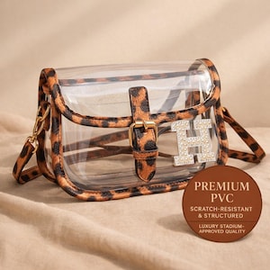 May include: Clear crossbody bag with leopard print trim and strap. The bag features a gold buckle and a rhinestone letter "H". The bag has a brown circular tag with the words "PREMIUM PVC" and "SCRATCH-RESISTANT & STRUCTURED".