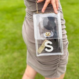 May include: A clear, rectangular crossbody bag with a gold chain strap. The bag contains sunglasses, a gold-colored item, and a large, sparkly letter "S". The bag is held against a background of green grass.