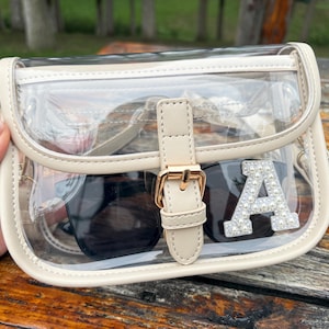 May include: A clear, rectangular handbag with a cream-colored trim and a gold buckle closure. The bag contains black sunglasses and a decorative letter "A" embellished with small, shiny beads. The bag is designed to be see-through, showcasing the contents.