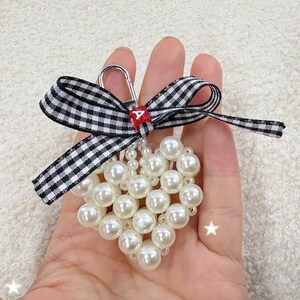 Può includere: Un portachiavi a forma di cuore realizzato con perle finte bianche. Presenta un fiocco a quadretti bianchi e neri e un fermaglio color argento. Un piccolo accento rosso con una lettera bianca è attaccato al fermaglio.