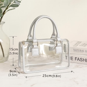 May include: A clear, rectangular handbag with silver-colored handles and trim. The bag measures 9.8 inches long, 5.9 inches high, and 3.5 inches wide. The bag has a zipper closure and is sitting on a marble surface.