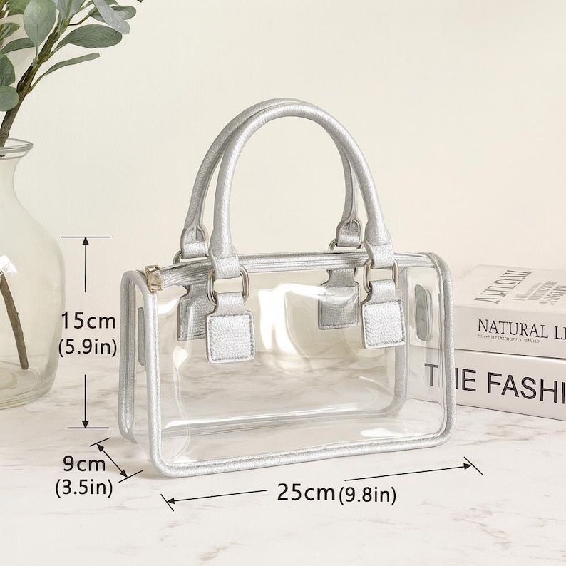 May include: A clear, rectangular handbag with silver-colored handles and trim. The bag measures 9.8 inches long, 5.9 inches high, and 3.5 inches wide. The bag has a zipper closure and is sitting on a marble surface.