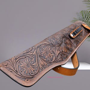 Puede incluir: Una funda de rifle de cuero marrón con intrincados grabados florales. La funda presenta un diseño curvo, una correa de cuero y un pequeño soporte para municiones. Los bordes están cosidos con hilo blanco, para un aspecto rústico y detallado.