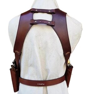 K&ouml;nnte beinhalten: Ein braunes Leder-Schulterholster mit zwei angebrachten Taschen und einem G&uuml;rtel. Das Holster verf&uuml;gt &uuml;ber verstellbare Riemen und Metallnieten. Das Design ist von hinten sichtbar und zeigt die Handwerkskunst und die Details.