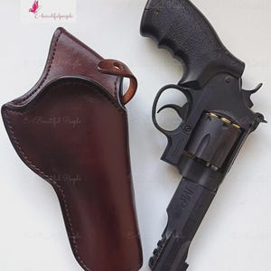Op de afbeelding: Een zwarte revolver met een getextureerde grip en een bruine leren holster. De holster is ontworpen om op het pistool te passen en heeft een lus voor een riem. Het pistool is geladen met kogels.