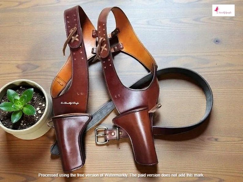 K&ouml;nnte beinhalten: Ein braunes Schulterholster aus Leder mit zwei Pistolenholstern, einem G&uuml;rtel und Riemen. Das Holster hat eine Schnalle und liegt auf einer Holzoberfl&auml;che. Eine kleine Topfpflanze befindet sich im Hintergrund.