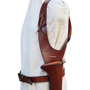 K&ouml;nnte beinhalten: Ein braunes Schulterholster aus Leder mit passendem G&uuml;rtel. Das Holster hat ein vertikales Design mit einer Tasche f&uuml;r eine Schusswaffe. Der Schulterriemen ist mit Nieten und einem dekorativen Lederband befestigt.