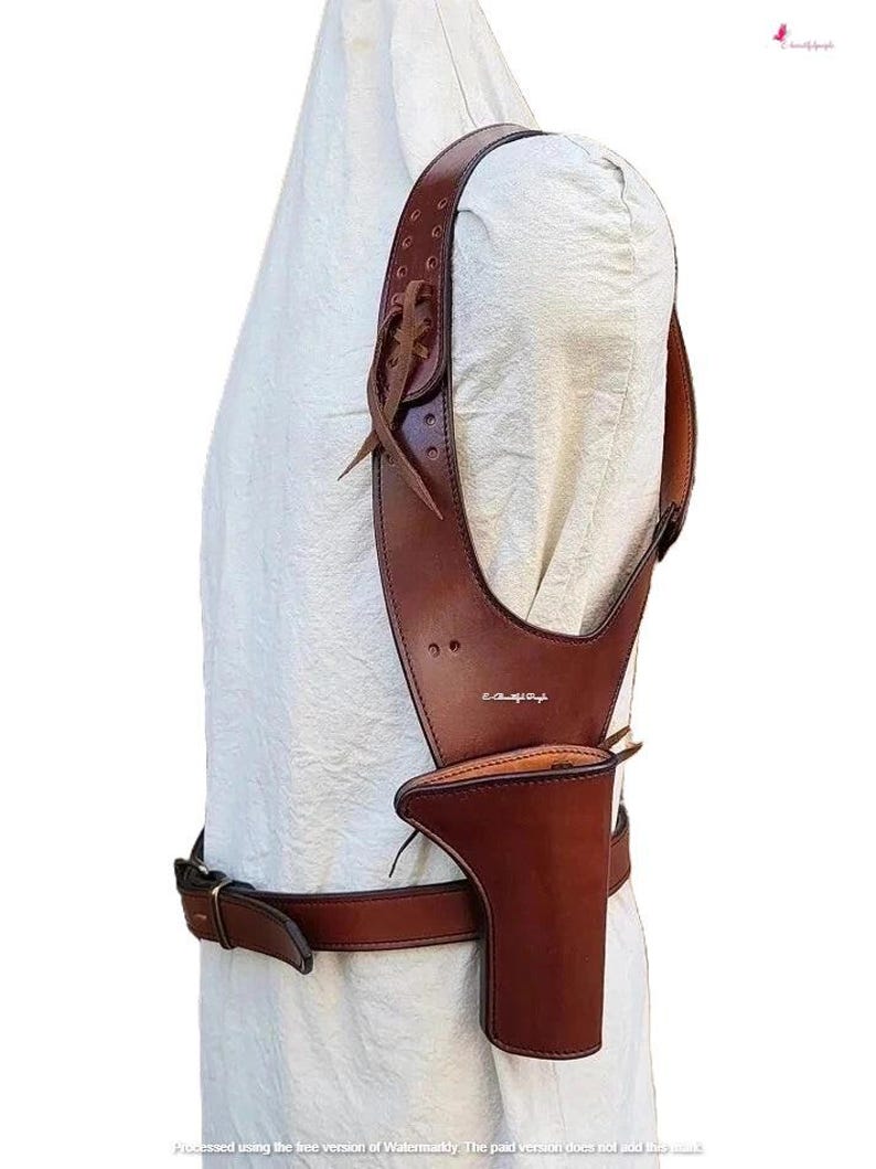 K&ouml;nnte beinhalten: Ein braunes Schulterholster aus Leder mit passendem G&uuml;rtel. Das Holster hat ein vertikales Design mit einer Tasche f&uuml;r eine Schusswaffe. Der Schulterriemen ist mit Nieten und einem dekorativen Lederband befestigt.
