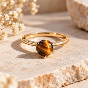 Op de afbeelding: Een gouden ring met een ronde, gepolijste tijgeroog edelsteen in een zeskantige zetting. De edelsteen vertoont karakteristieke bruine en gouden banden met een chatoyante glans, gemonteerd op een gladde, gepolijste gouden ring.