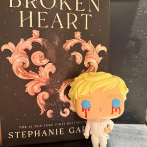 May include: A Funko Pop figure of a character with blonde hair and white clothing, with red tears streaming from its blue eyes. The figure is positioned in front of a book titled "Once Upon a Broken Heart" by Stephanie Garber.